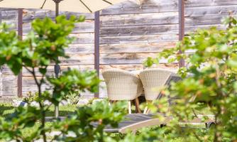 Two comfortable chairs are placed in the garden under a sunshade. In the background, a wooden-clad wall can be seen.