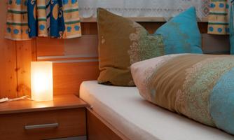 A cozy bedroom with colorful cushions and a bedside table. A soft lamp provides warm light.