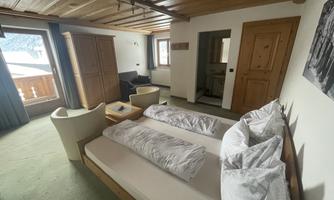 A cozy room with a wooden ceiling and two beds. There is a small living area with comfortable armchairs and access to a balcony.