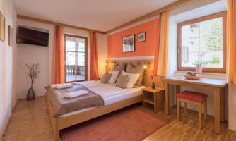 A cozy bedroom with a large bed and orange accents. There is a desk, a chair, and a window with a view of the outdoors.