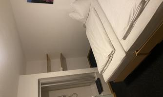 A simple bedroom with two single beds and white bedcovers. In the background, there is a bathroom with a shower and a small shelf.