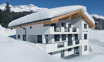 A modern building in the snow with a snow-covered roof. Behind the house, mountains and coniferous trees extend.