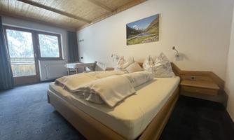 A cozy bedroom with a double bed and wooden beam ceiling. A table and chairs are also available, as well as a picture of a landscape on the wall.