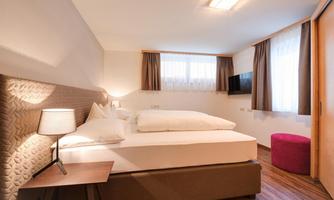 A modern bedroom with a double bed and a nightstand.  
Large windows let in plenty of light, and there is a small pink stool.