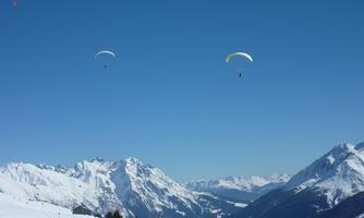 Two paragliders float over snow-covered mountains. The clear, blue sky complements the wintry landscape.