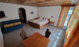 A cozy room with a double bed and a sofa. Large windows with bright curtains provide plenty of natural light.