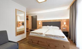 A modern room with two wooden single beds and a large window front. The decor is simple and friendly, featuring a cozy armchair and a mirror.
