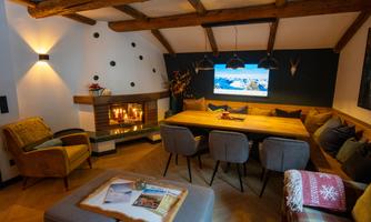 A cozy living room with a wooden beam ceiling and a large dining table. In the background, a fireplace and a television can be seen.