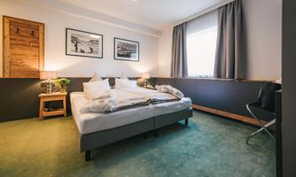 A modern bedroom with a spacious double bed and green carpet. The walls are decorated with photos, and there are curtains that let in daylight.