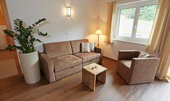 A cozy living area with a beige sofa, an armchair, and a small table. Some plants are growing by a window and there is warm light from a lamp.