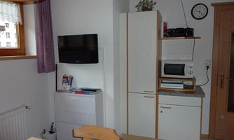A cozy kitchen with a table, a television, and a refrigerator.  
On the wall, there is a microwave and above it a shelf with a radio.
