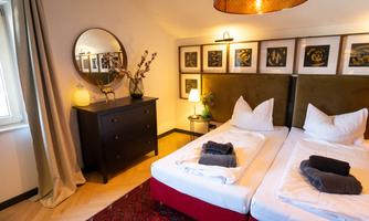 A cozy bedroom with two single beds and stylish decorations. The room features a carpet, a mirror, and a dresser.