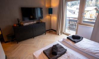 A modern bedroom with two beds and a television. The room has windows with a view of the mountains and is decorated in a welcoming style.