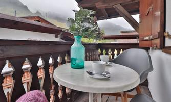 A cozy balcony with a round table and two chairs. On the table sits a glass vase with green plants and two coffee cups.