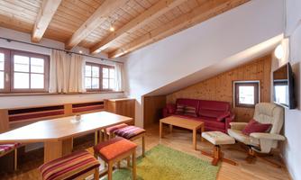 A cozy living room with wooden ceilings and modern furnishings. The room features a dining table, a seating area, and large windows that let in plenty of light.