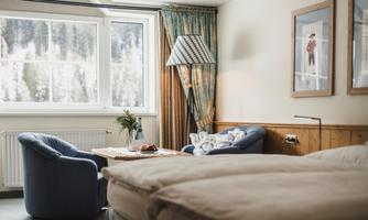 A cozy hotel room with two blue armchairs and a table. The window lets in plenty of daylight and offers a view of nature.
