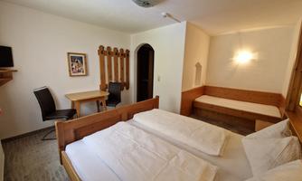 A bright room with a double bed and a single bed. There is a table with a chair and a wall with wooden shelves.
