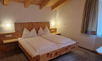 A cozy bedroom with a wooden double bed. The walls are light, and there are windows with curtains that let in daylight.