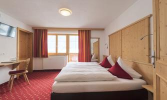A cozy hotel room with two beds and red cushions. Large windows let in plenty of light.