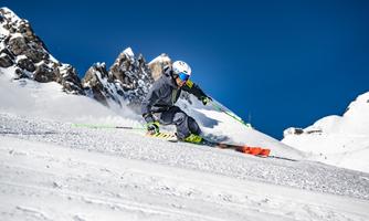 A skier glides quickly over the snowy slope. In the background, the snow-covered mountains and the clear blue sky can be seen.