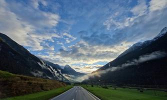 A picturesque road winds through a green landscape with mountains in the background. The sky is cloudy and there is a hint of fog.