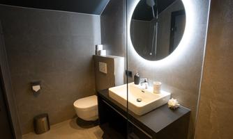 A modern bathroom with dark walls and a round mirror. The fittings include a sink, a toilet, and practical accessories.