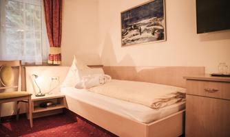A simple room with a single bed and a desk. A painting hangs on the wall, and there is a chair as well as a lamp.