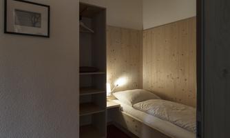 A simple bedroom with a single bed and wooden paneling. The room has warm lighting and a cozy atmosphere.