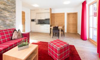 A modern living room with a red carpet and a comfortable couch. The open kitchen and dining area are well lit and brightly furnished.