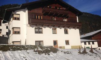 A cozy, multi-story house in alpine style, surrounded by snow. The façade is white with wooden elements, and mountains can be seen in the background.