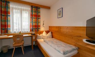 A simple bedroom with a single bed and a desk. The walls are clad in wood and the windows have colorful curtains.