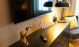 A stylish living room with a dark dresser, a large television, and a lamp. A welcome sign and decorative elements create an inviting atmosphere.