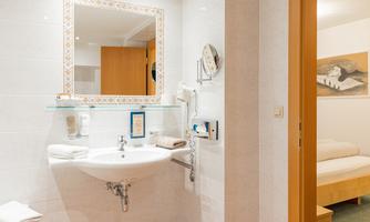 A modern bathroom with a sink and a mirror. In the background, a cozy bedroom is visible.