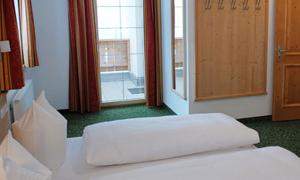A simple bedroom with two beds and white bedcovers. The windows have curtains and there is a wooden hook for the clothes.