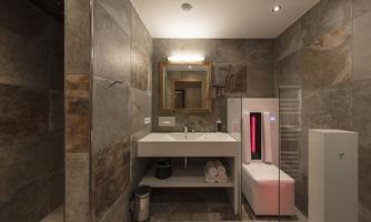 A modern bathroom with an elegant sink and a large mirror. The walls are clad with stone tiles, and a refrigerator is situated next to the sink.