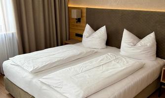 A cozy bedroom with a large double bed and white bedding. The room has a warm atmosphere with a window and blackout curtains.
