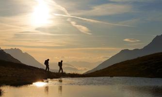 Two hikers walk along a path at sunset. In the background, there are mountains and a calm lake.