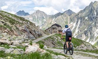 A cyclist rides on a gravel path in the mountains. In the background, impressive peaks and an alpine hut can be seen.