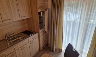 A modern kitchen with wooden cabinets and a stainless steel sink. In the background, there is a refrigerator and a table with a chair.