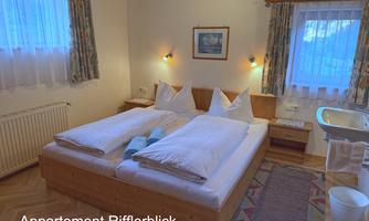 A cozy bedroom with two beds, light curtains, and soft lighting. The room features a small table and a sink.