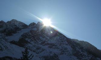 A steep mountain range with snow-covered peaks. The sun shines brightly over the mountains, creating a radiant light effect.