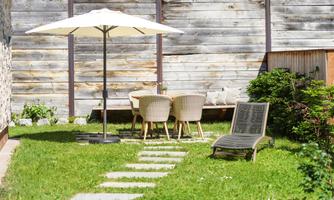 A cozy garden with a sunshade and a table with chairs. The lawn is green and well-maintained, with a deck chair and stone slabs leading to the seating area.