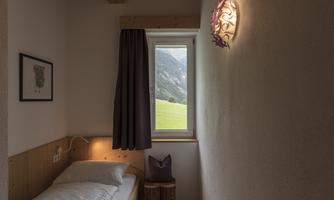 A simple and cozy room with a single bed and wooden details. The window offers a view of the green landscape and the mountains.