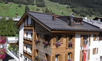 A cozy hotel with a rustic wooden design and windows adorned with flower boxes. In the background, green mountains can be seen.