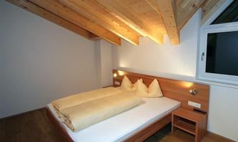 A cozy bedroom with a double bed and wooden ceiling. There is a window that lets light in, and a small bedside lamp.