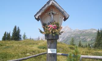 A wooden cross on a post with colorful flowers is placed in a green, mountainous landscape. In the background, majestic mountains and a clear blue sky are visible.