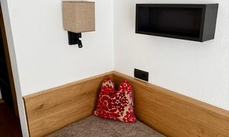A cozy bedroom with a simple bed and a red decorative pillow. On the wall, there is a stylish lamp and a shelf.