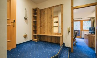 An inviting hallway with wooden shelves and a blue carpet area. In the background, windows with a view and a workspace are visible.