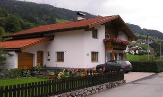 A cozy house with a red gabled roof and flower balcony. It is situated in a picturesque landscape with green hills in the background.