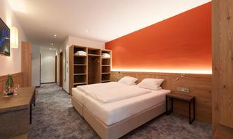 A modern hotel room with a double bed and a warm, orange accent wall. The room is well-lit and features a wardrobe as well as stylish furnishings.